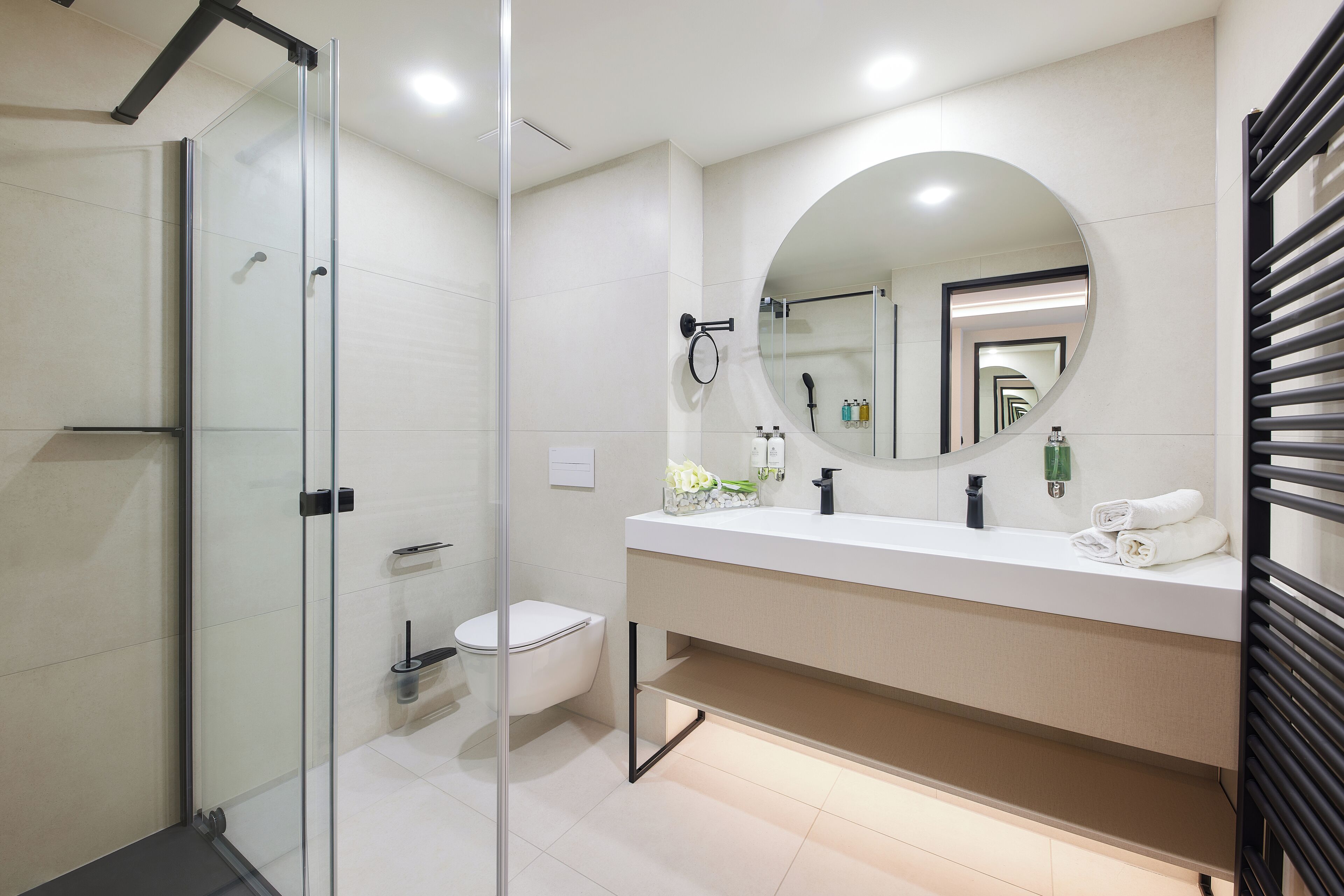 Ein modernes Badezimmer mit einer Duschkabine aus Glas, einer Toilette, einem weißen Waschbecken mit rundem Spiegel und einem Handtuchhalter.