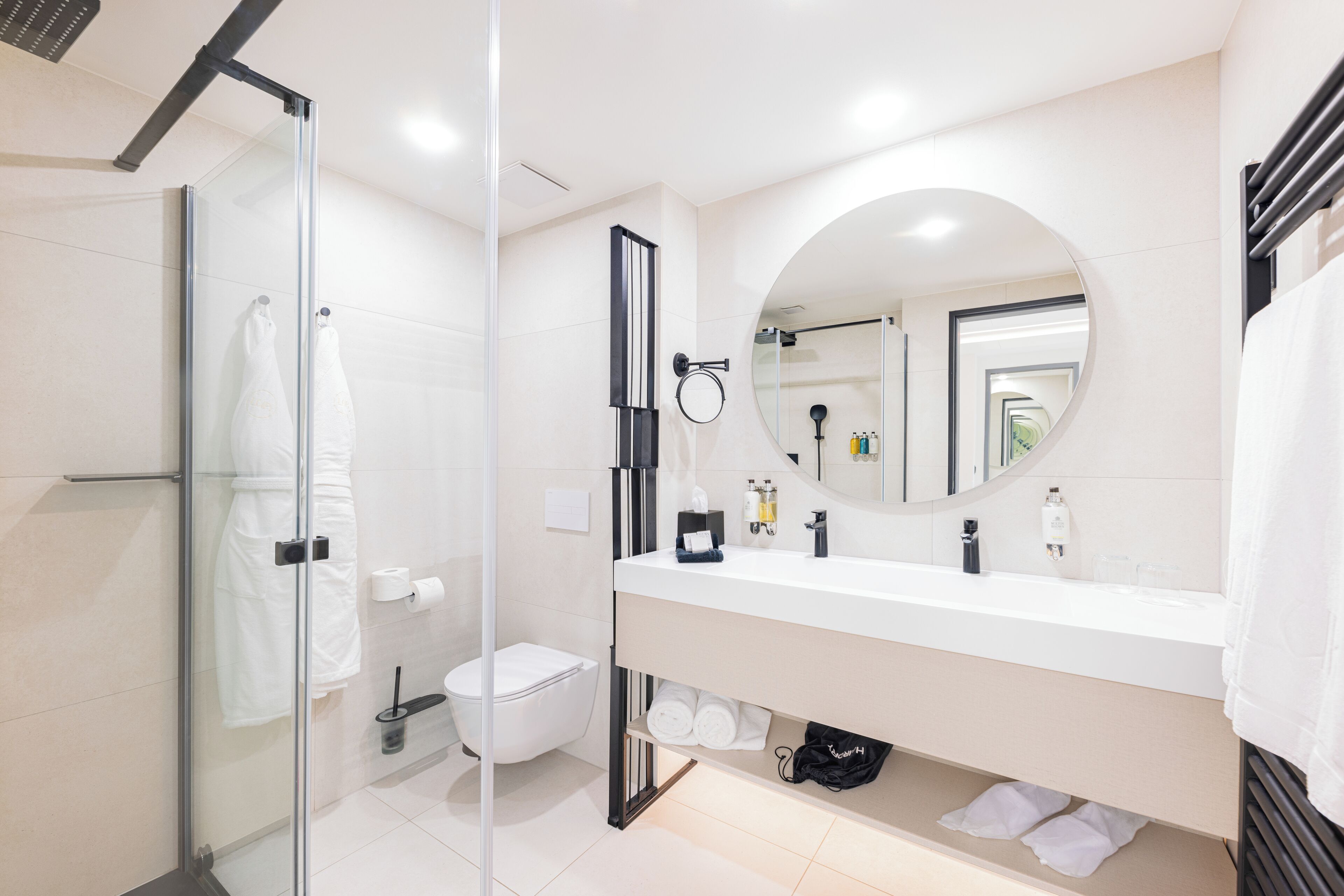 A modern bathroom with a glass shower enclosure, a white sink with a round mirror, and neatly arranged toiletries.