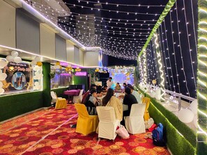 A decorated indoor event space with tables and chairs, string lights, and a stage area. - Hotel Jagat inn (Haridwar)