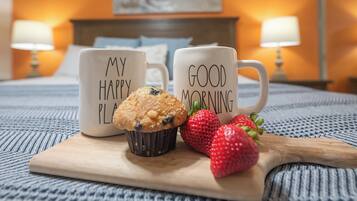 A bed with two white mugs on a wooden tray, one with "MY HAPPY PLACE" and the other with "GOOD MORNING". There are two strawberries and a muffin on the tray.