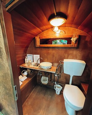 A compact, wooden bathroom with a sink, toilet, and a small shelf above the mirror.