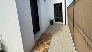 Un étroit couloir extérieur doté d’un plancher en bois, d’une porte noire et d’une plante en pot.