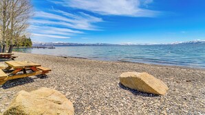 A pebble beach with picnic tables and a large rock, overlooking a calm lake with mountains in the distance.