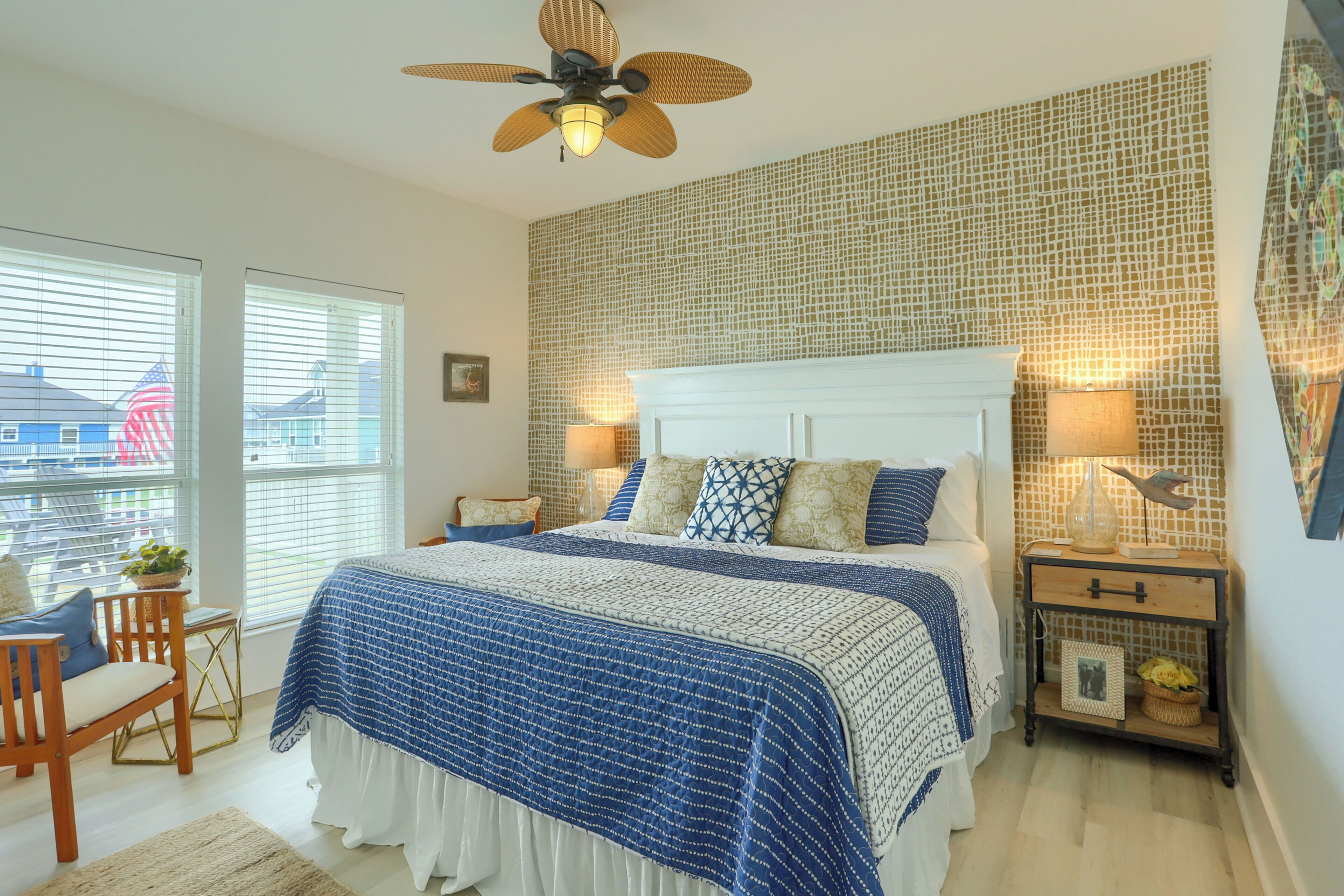 A bedroom with a large bed, a ceiling fan, a nightstand with a lamp, and a view of the outside through the window.