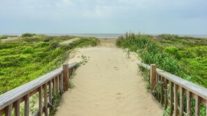 Un sentier de plage sablonneux, bordé de rambardes en bois, qui mène à la mer.