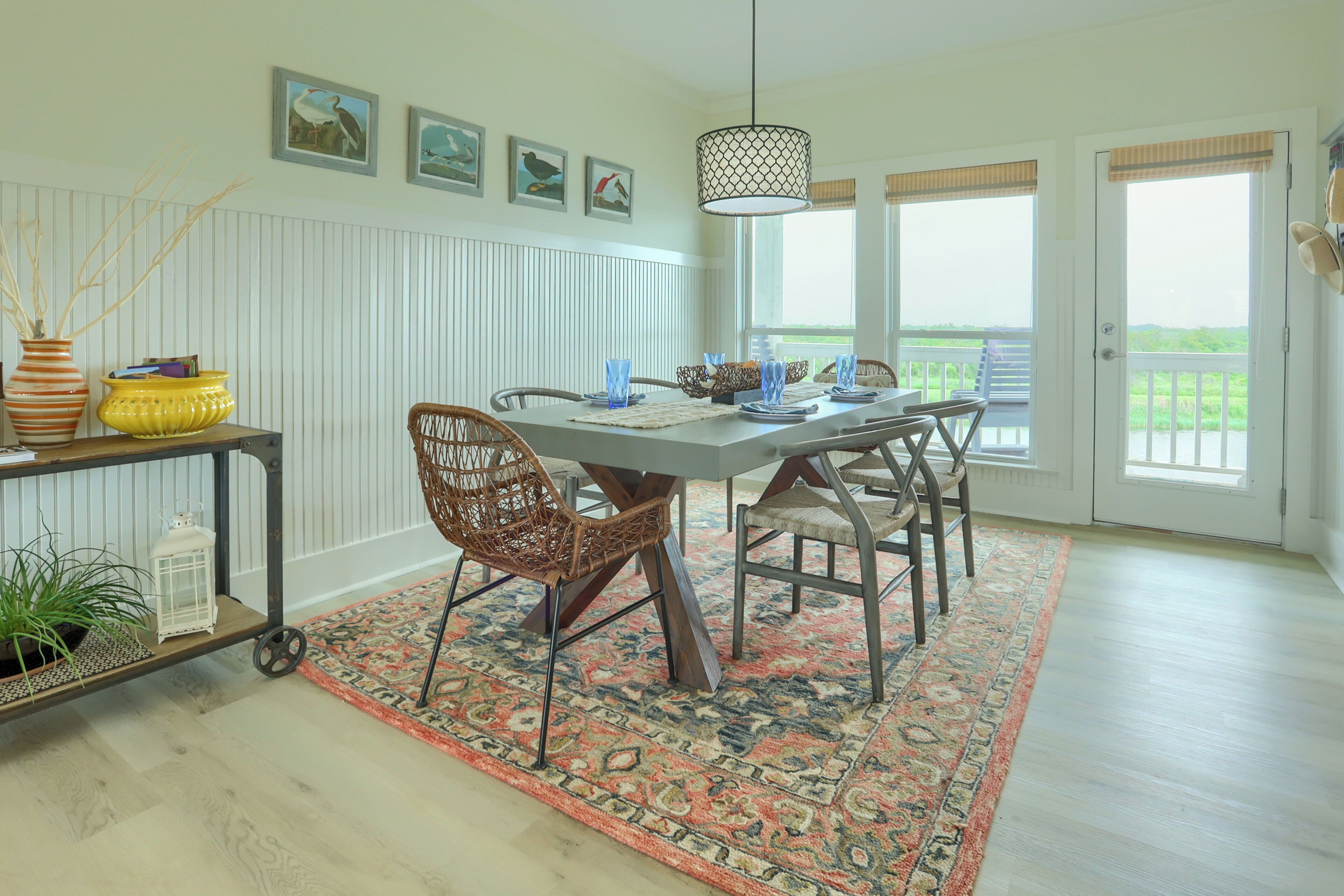 Een eetruimte met een tafel gedekt voor vier personen, omringd door rieten stoelen, een vloerkleed met een patroon en een hanglamp.