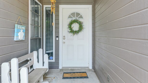 A front porch with a white bench, a welcome sign, a door with a wreath, and a doormat.