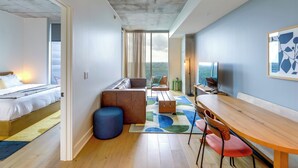 Quarto de hotel moderno com uma cama, área de estar com mesa e cadeiras, um sofá e uma janela grande com vista.