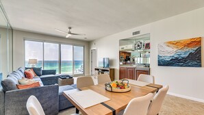 A modern living room with a sofa, dining table, and chairs, a kitchen area with cabinets and appliances, and a large window with a view of the ocean. - Oceanfront Destin Condo With Pool & Beach Access (Destin)