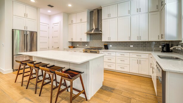 A modern kitchen with white cabinetry, a stainless steel refrigerator, a large island with bar stools, and a wood floor. - Spacious Austin Home: Near S Congress & Downtown! (Austin)