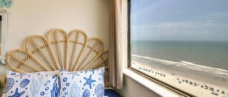 A room with a bed, a window with a view of the beach, and a wall with a decorative item.