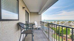 A balcony with a table and chairs, a view of the ocean, and a parking lot in the background.