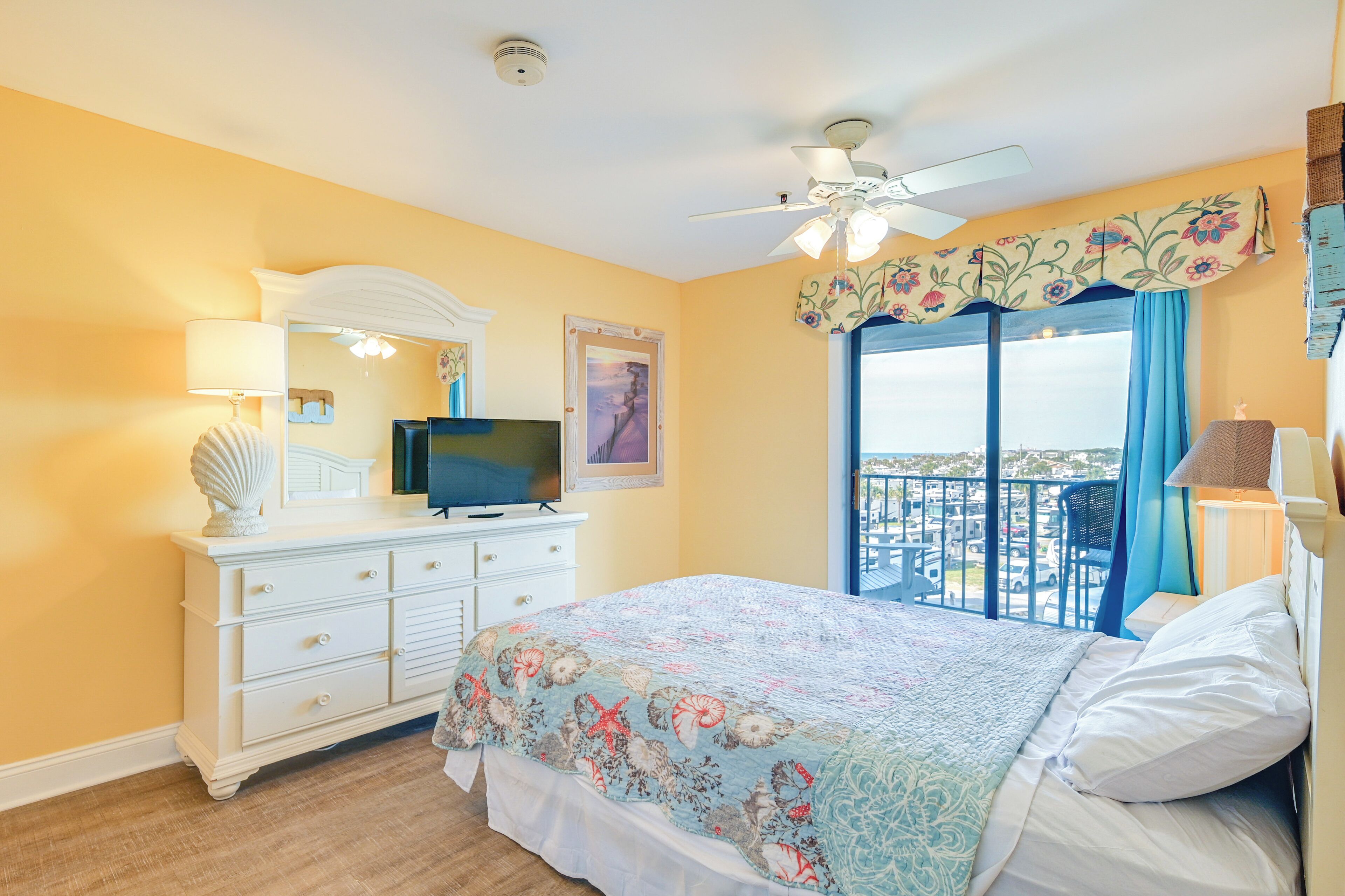 Une chambre à coucher comprenant un lit, une commode, un téléviseur, un ventilateur de plafond et un balcon avec vue sur la ville.
