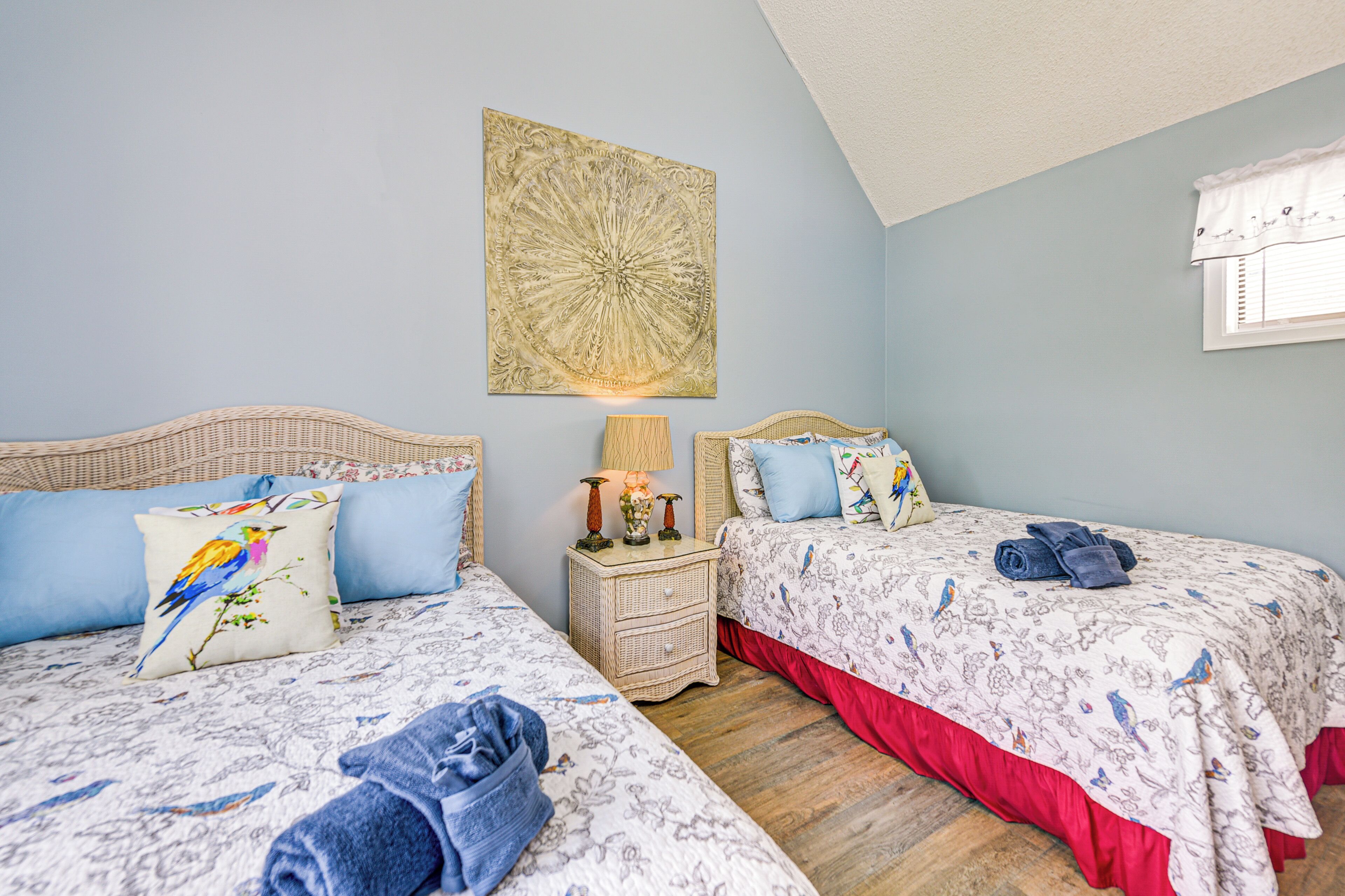 Ein Schlafzimmer mit zwei Betten, jeweils mit Blumenmuster bezogen und blauen Kissen, einem Nachttisch mit Lampe sowie einem Wandbild.