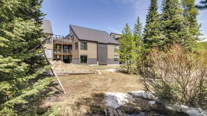 Une maison moderne à deux étages, dotée d’un toit en métal, entourée d’une forêt dense de conifères et traversée par un petit ruisseau.
