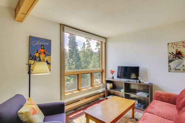 A living room with a large window, a sofa, a coffee table, a TV, and a bookshelf.