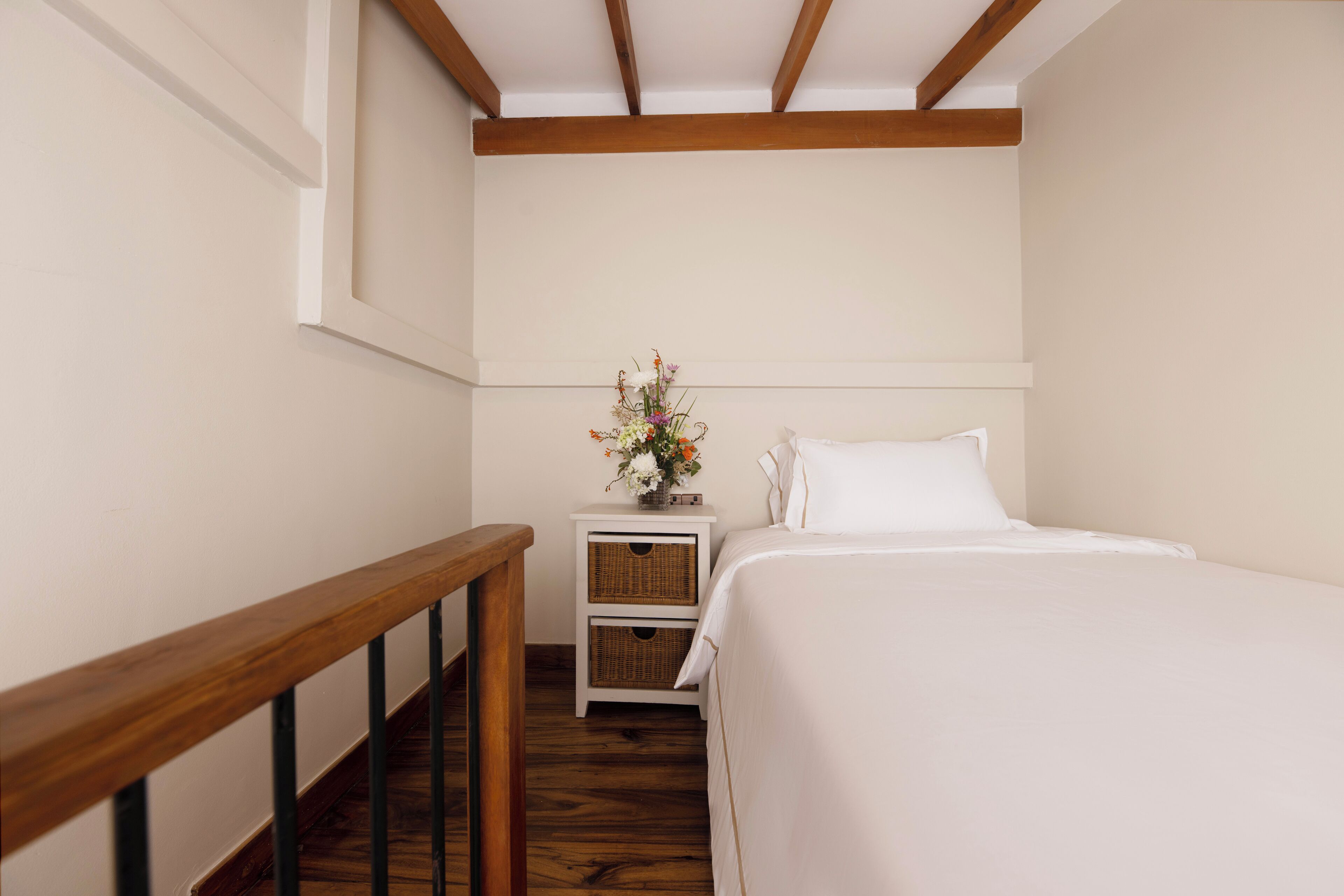A bedroom with a wooden staircase, a white bed, a nightstand with a vase of flowers, and a white wall.