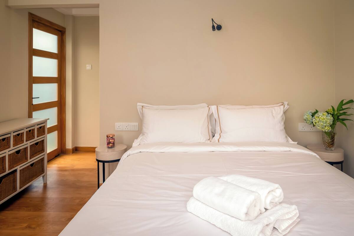 Uma cama bem arrumada com lençóis brancos, uma mesa de cabeceira com um vaso de flores e um aparador de madeira com cestos.