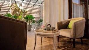 A cozy room with a wooden floor, a beige armchair with a green pillow, a small round table with a tray of items, and potted plants. - Ara by Colombo Villas  (Colombo)