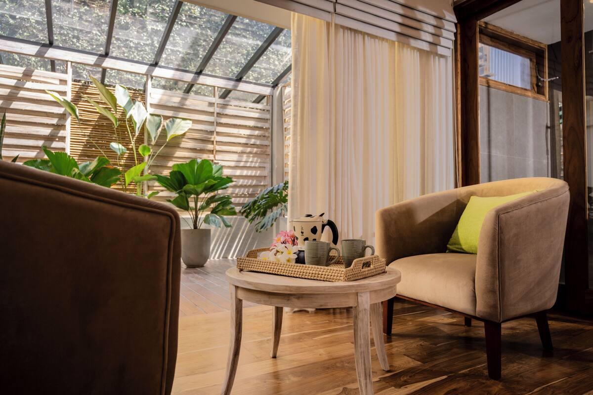 Um quarto acolhedor com piso de madeira, uma poltrona bege com almofada verde, uma pequena mesa redonda com uma bandeja de objetos e plantas em vasos.