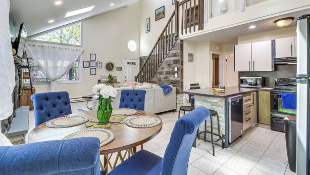 A spacious living area with a dining table, blue upholstered chairs, a kitchen with modern appliances, and a staircase leading to an upper level. - Hot Tub & Lake Beach Access: Tobyhanna Retreat! (Tobyhanna)
