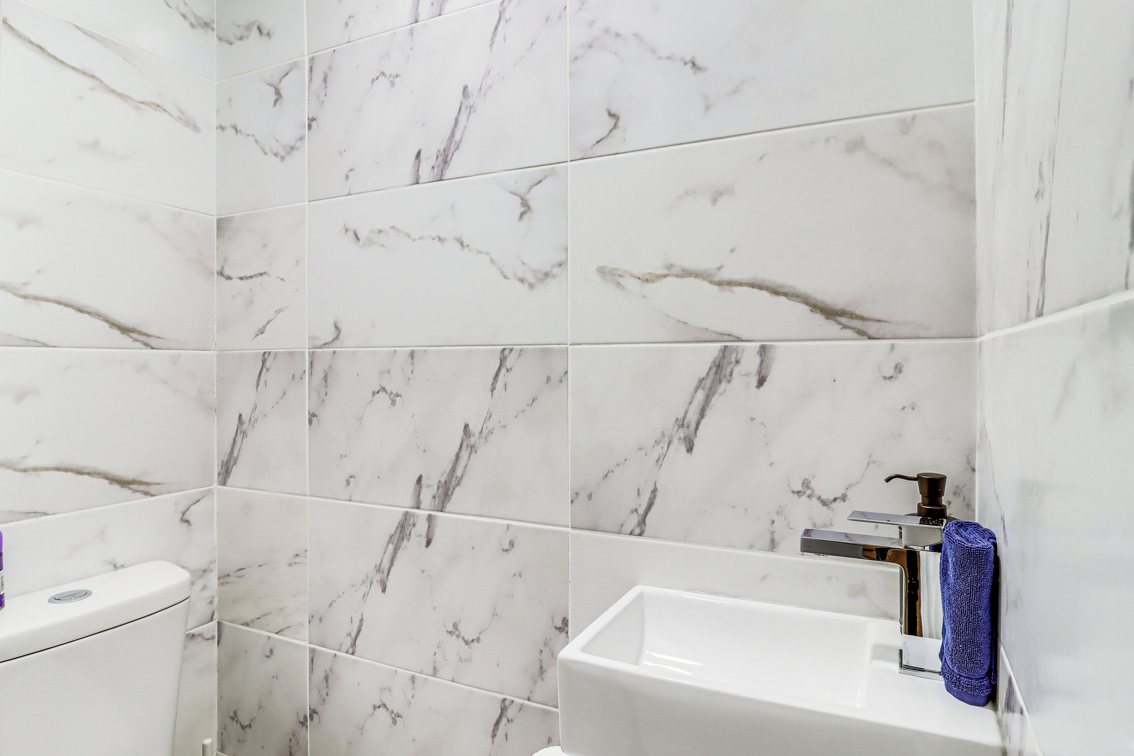 Un baño con baldosas de mármol blanco, un lavabo blanco y un dispensador de jabón.