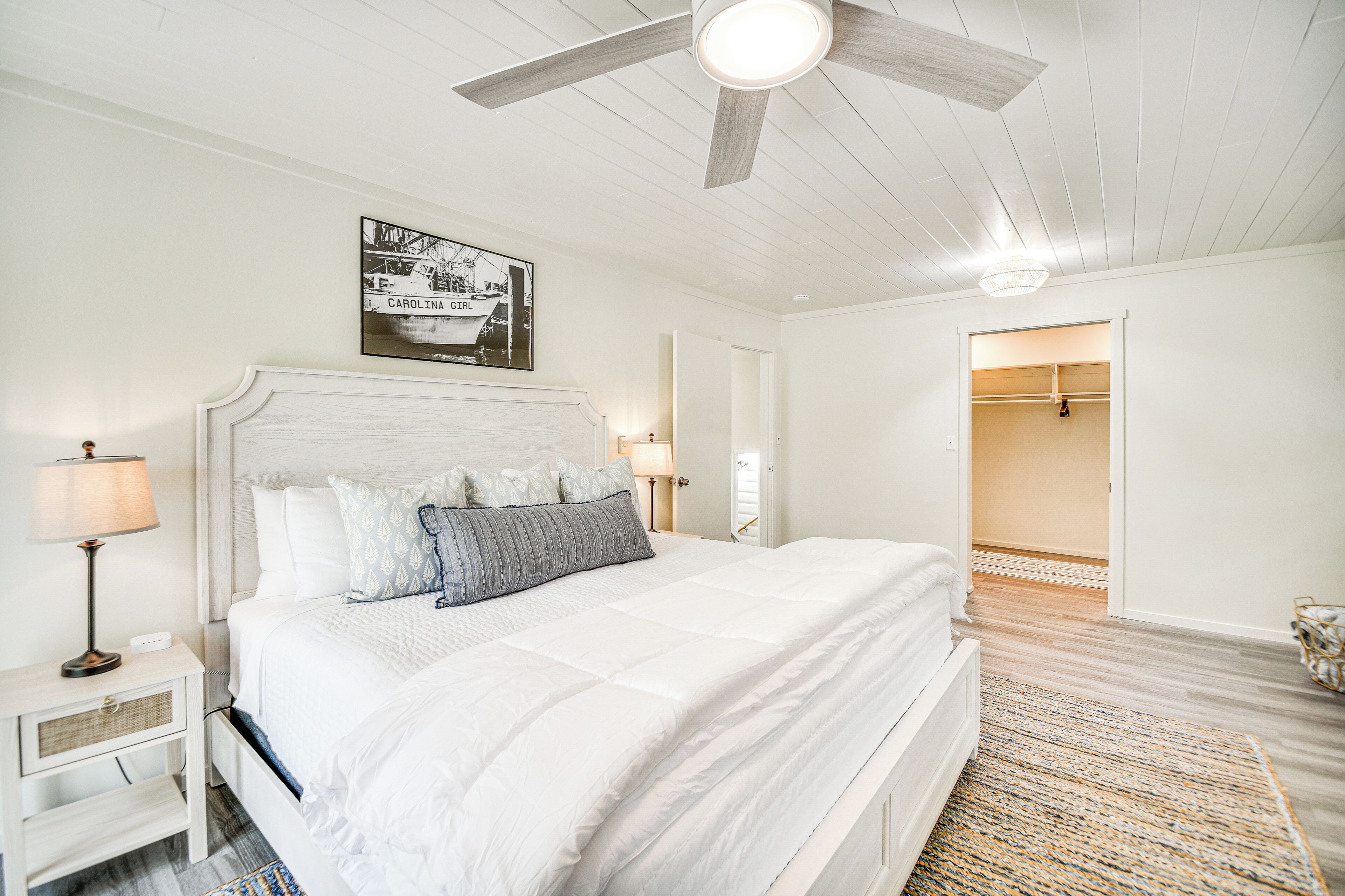 A bedroom with a large bed, a ceiling fan, a nightstand, a lamp, and a framed picture on the wall.