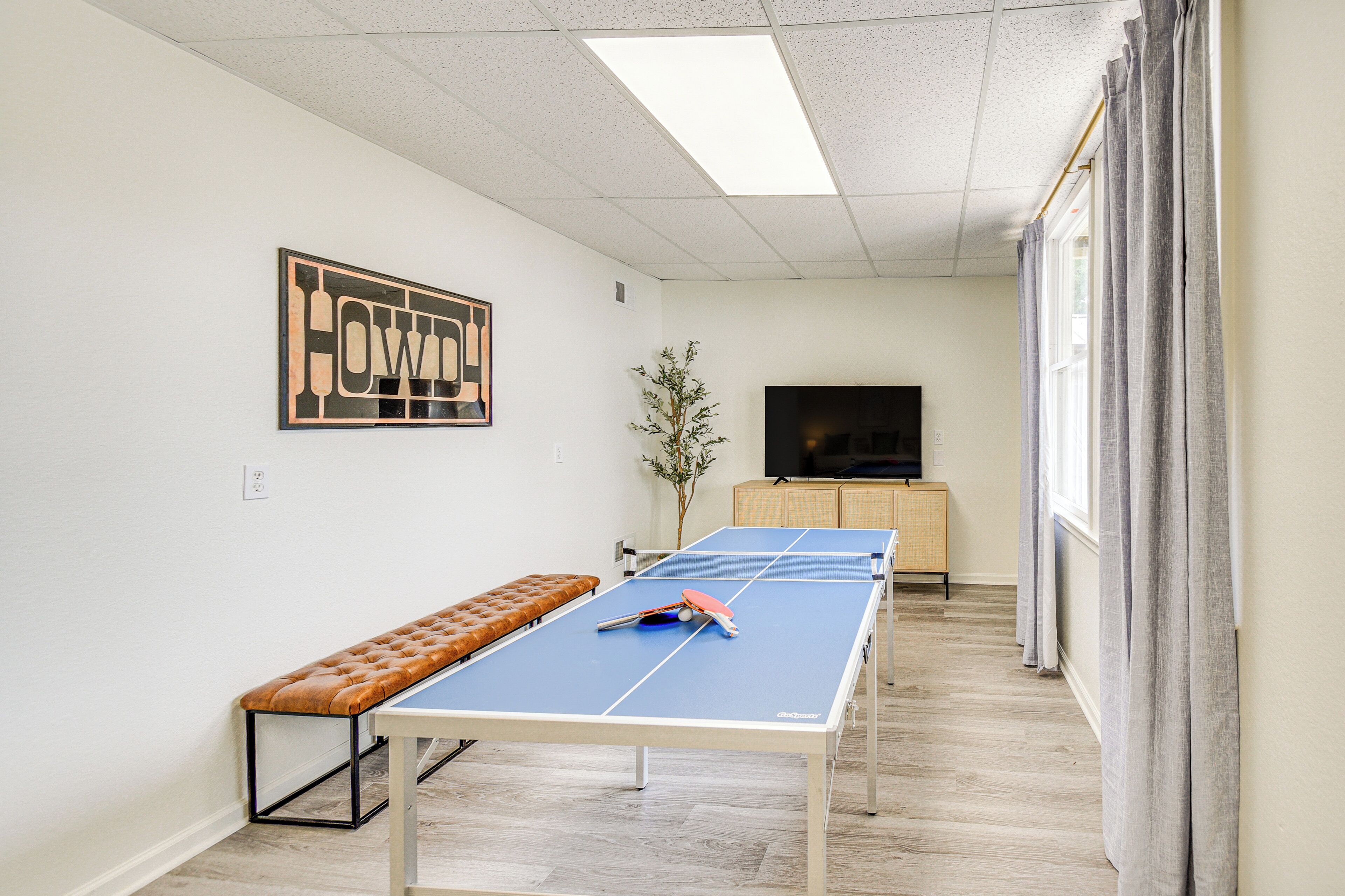 A ping pong table with a bench, a television, and a cabinet in a room with a light-colored floor and walls.