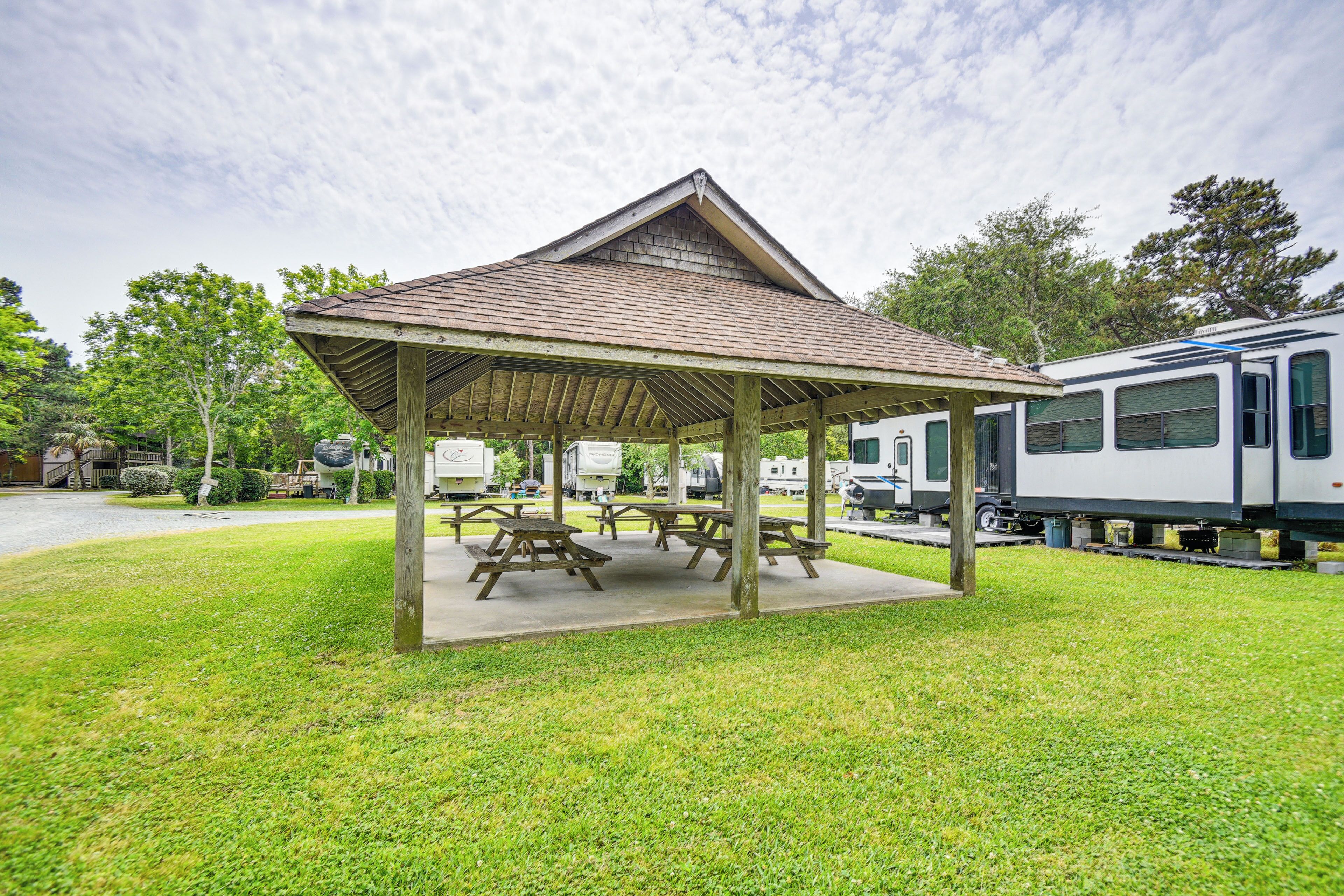 A covered picnic area with benches and picnic tables, surrounded by grass and trees.