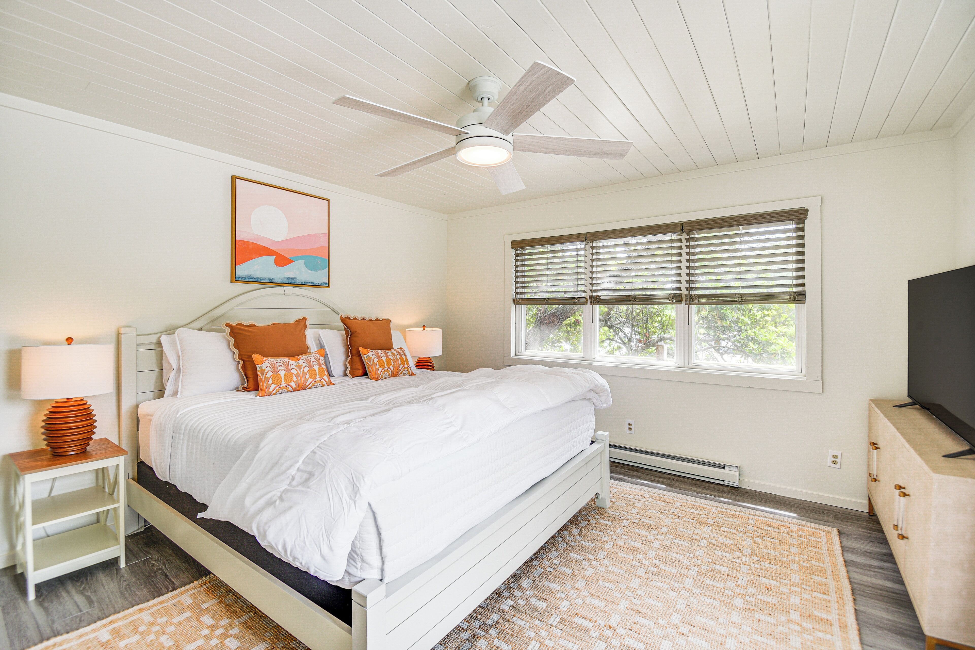 A bedroom with a bed, a ceiling fan, a window with blinds, a bedside table with a lamp, and a TV stand.
