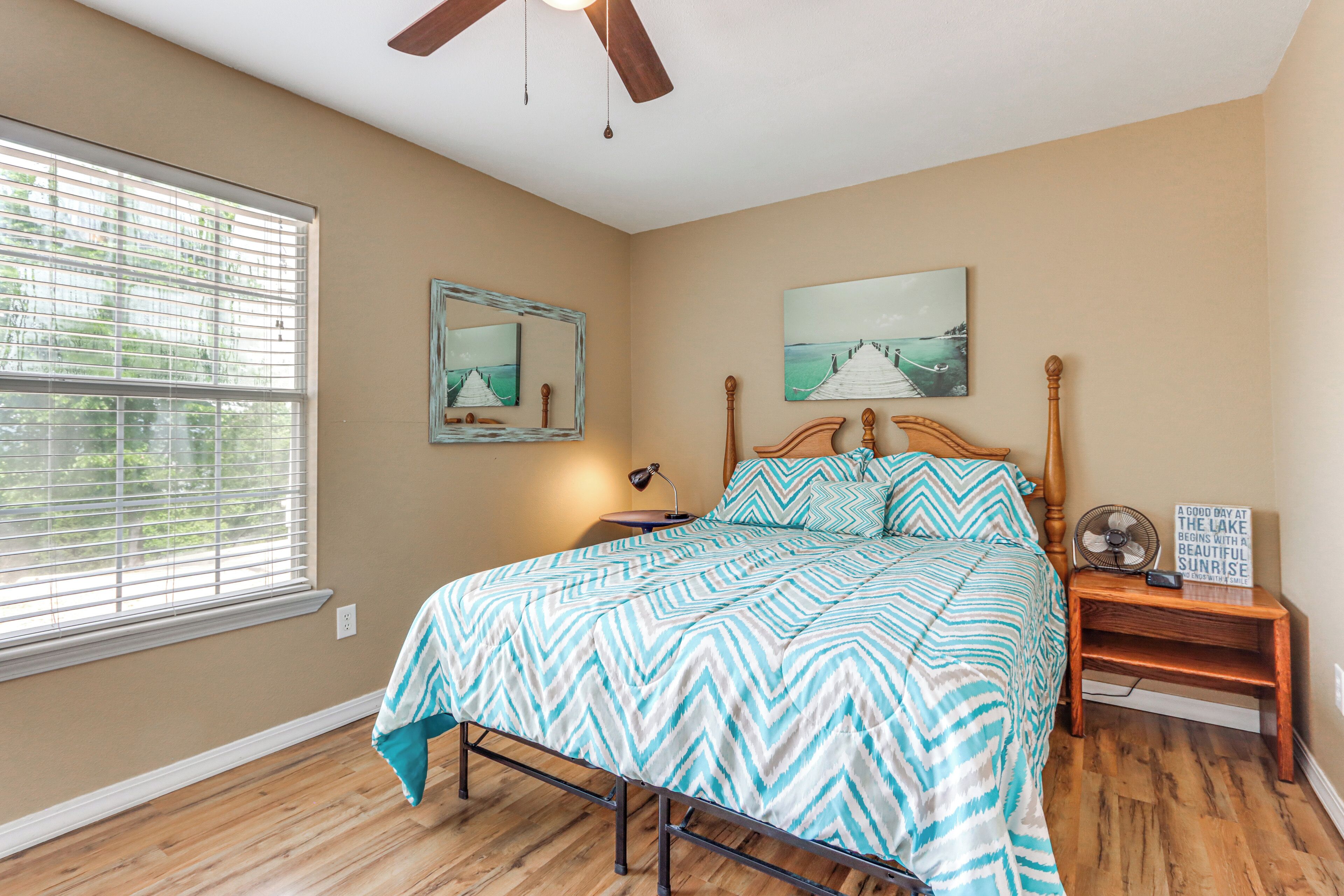 A bedroom with a bed, a nightstand, a ceiling fan, a window with blinds, and artwork on the walls.