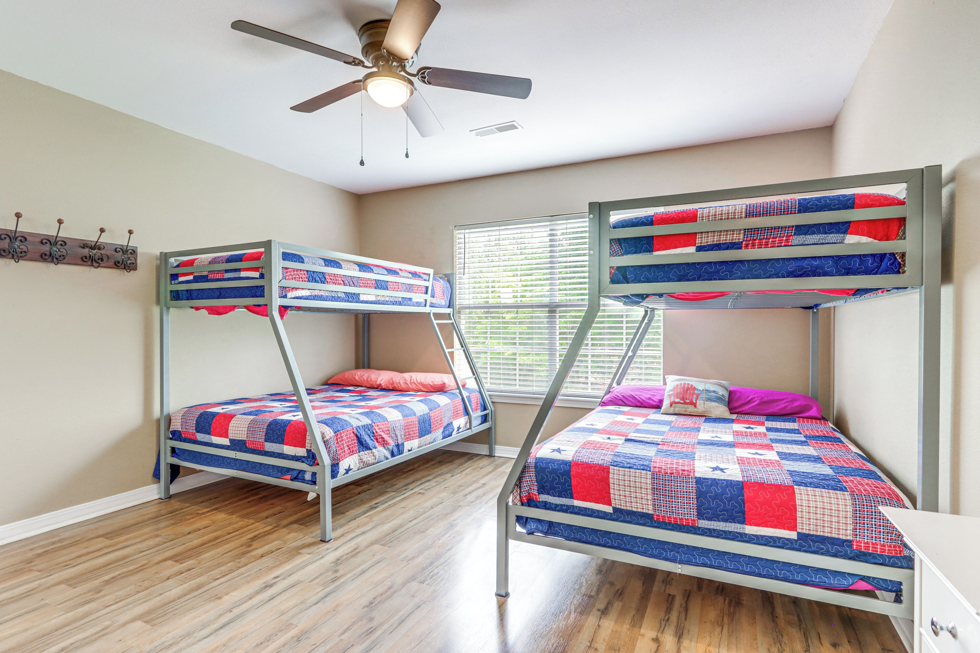 A room with two bunk beds, wooden flooring, a ceiling fan, and a window with blinds.