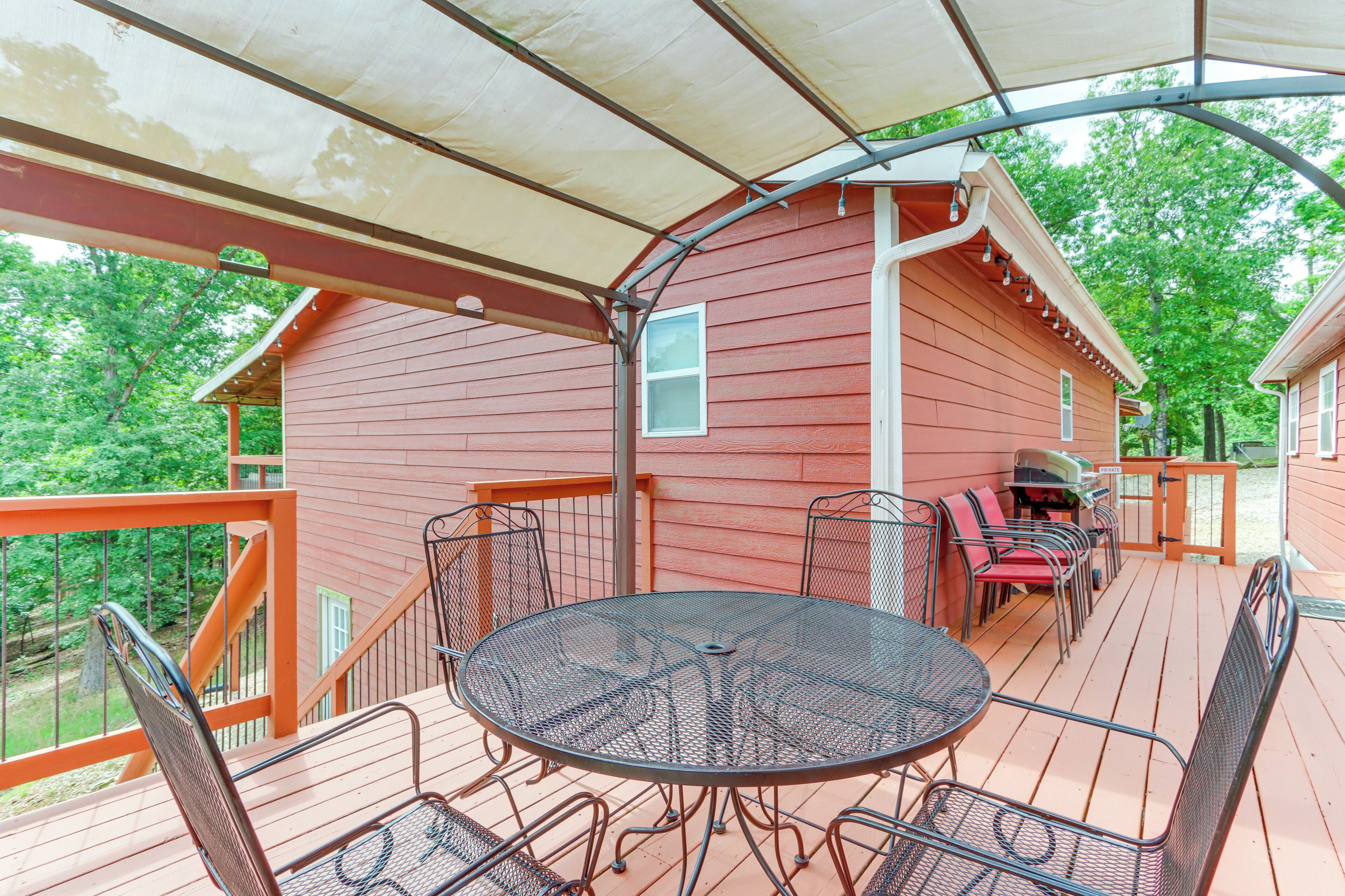 A covered outdoor deck with a metal table and chairs, a barbecue grill, and a wooden railing.