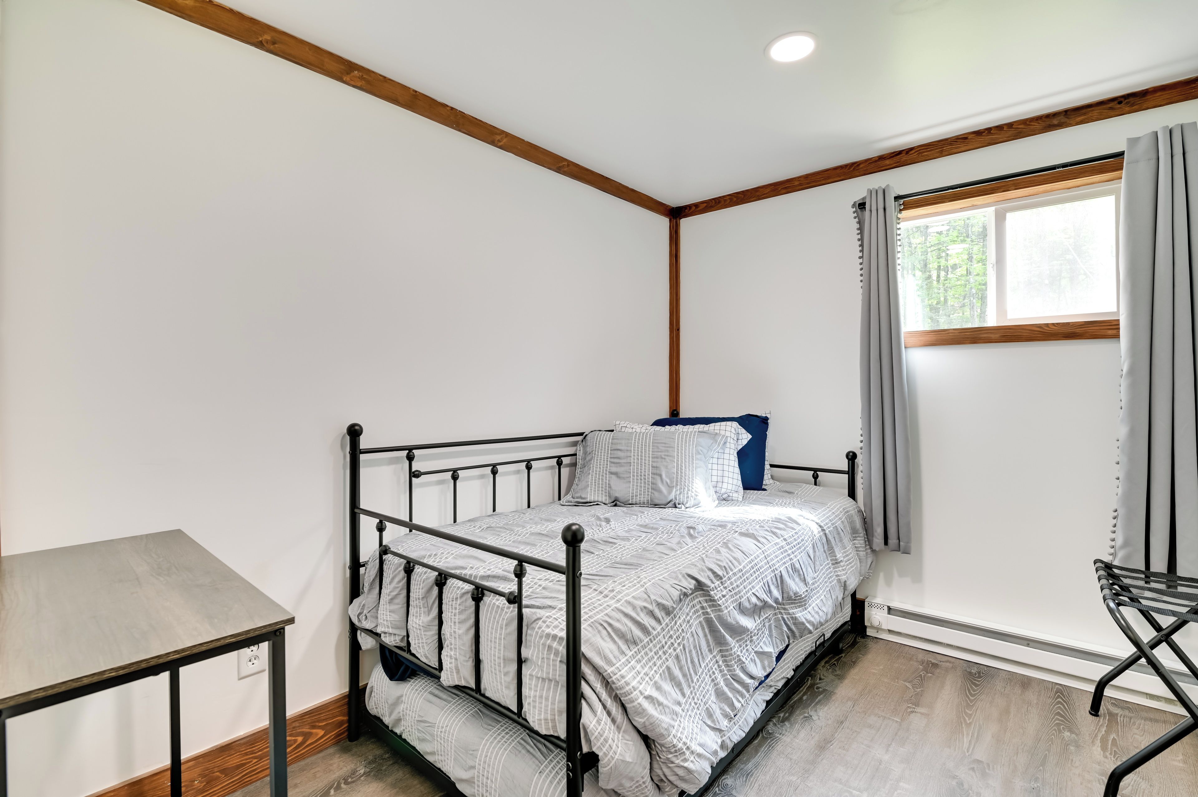 A small, single-bed room with a metal frame, a wooden table, and a window with curtains.