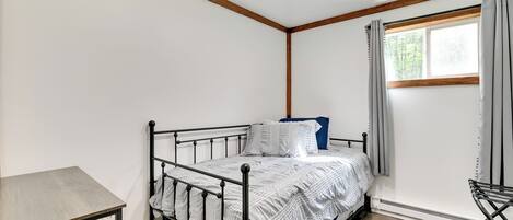 A small, single-bed room with a metal frame, a wooden table, and a window with curtains.