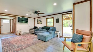 A spacious living room with a large sectional sofa, a leather armchair, and a small side table. A ceiling fan is visible, and there is a large window providing natural light. - Cozy Catskills Cabin - 12 Mi to Windham Mountain! (East Durham)