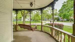 Een veranda met een houten schommel, een lamp en uitzicht op een woonstraat met huizen en groen.