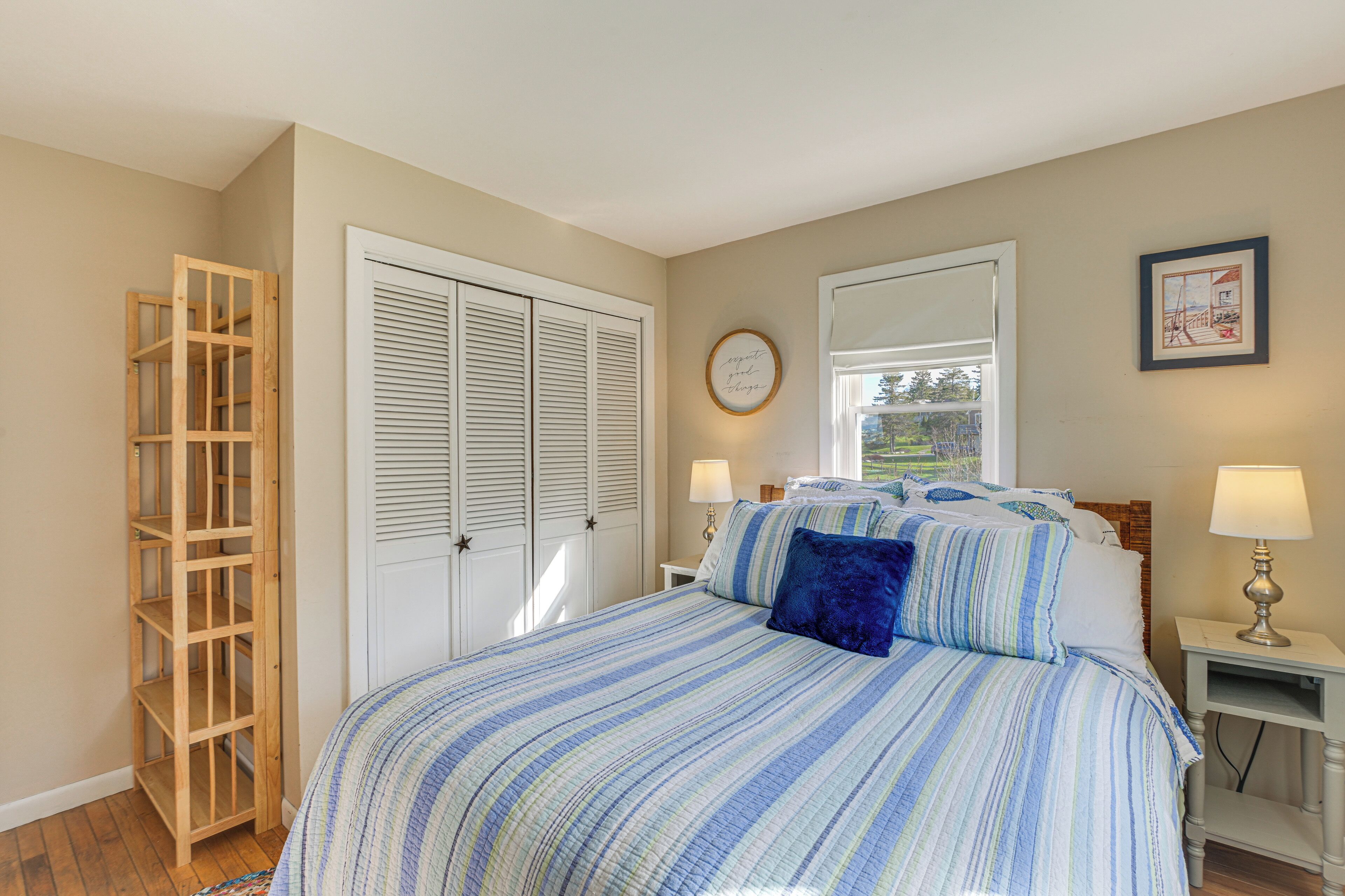 A bedroom with a bed, bedside tables, a wooden shelving unit, a wardrobe, and a window with blinds.
