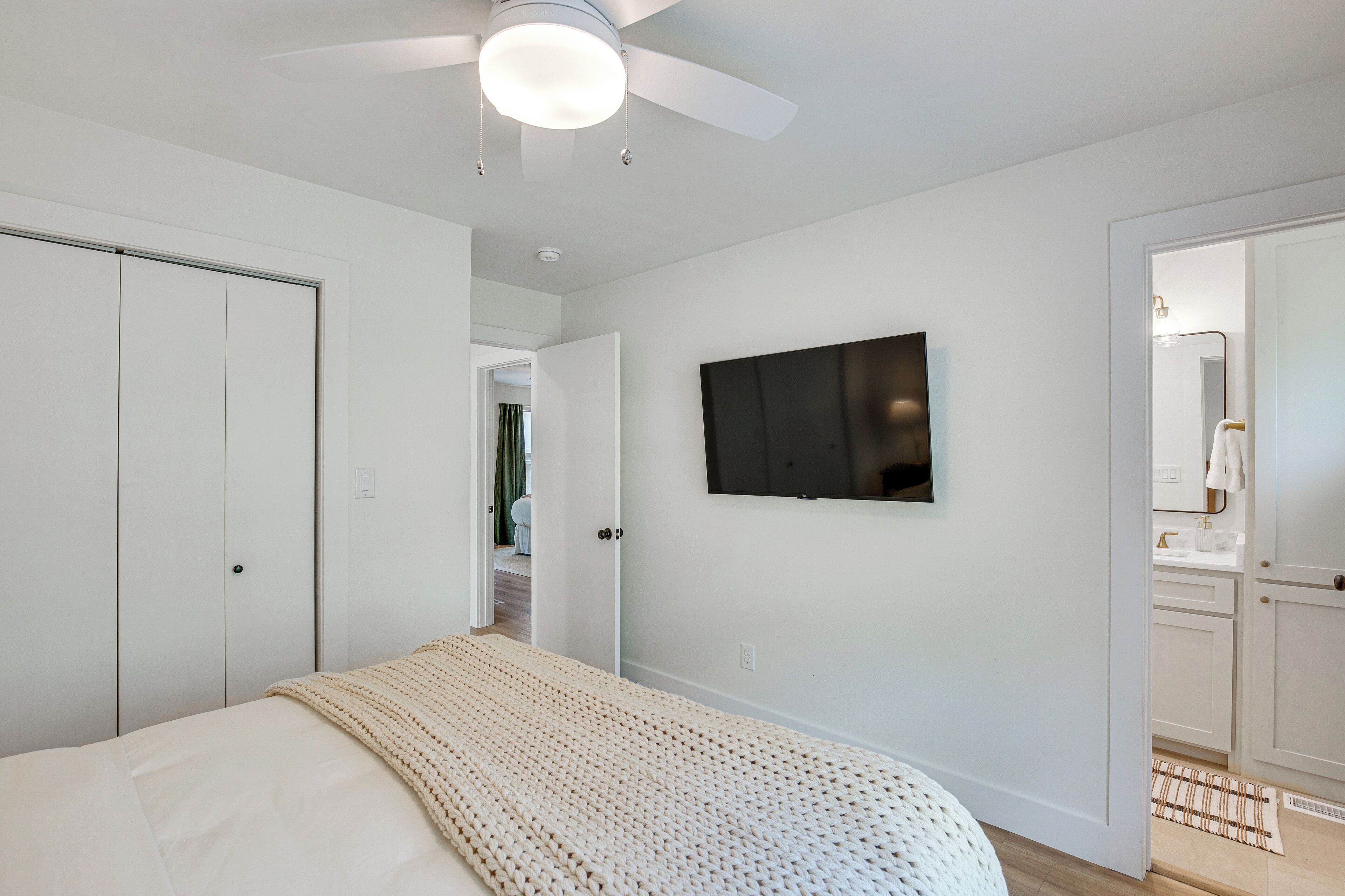 A bedroom with a bed, a ceiling fan, a TV mounted on the wall, and a bathroom visible through an open door.