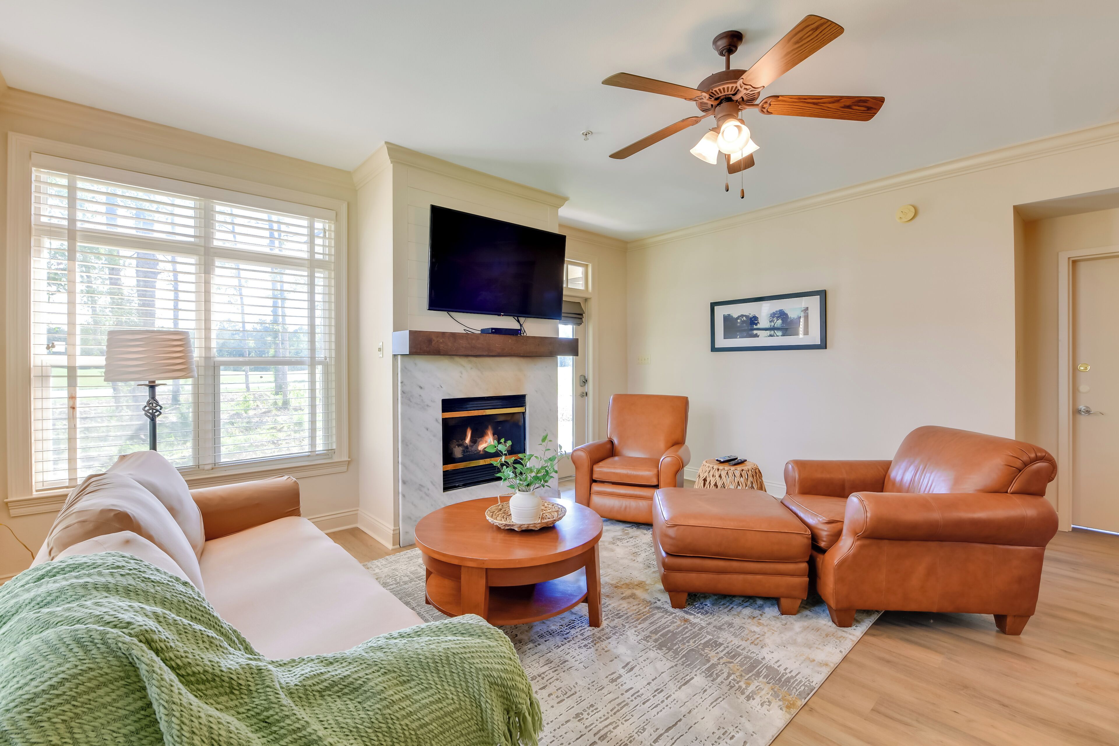 A living room with a fireplace, leather furniture, a ceiling fan, and a large window with blinds.