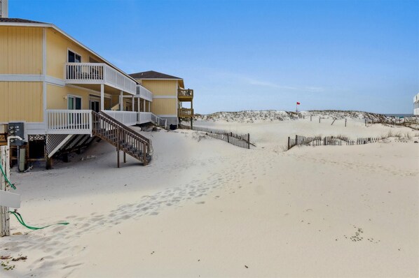 A beachfront hotel with a staircase leading to a balcony, a sandy beach in the background, and a red flag on a pole.