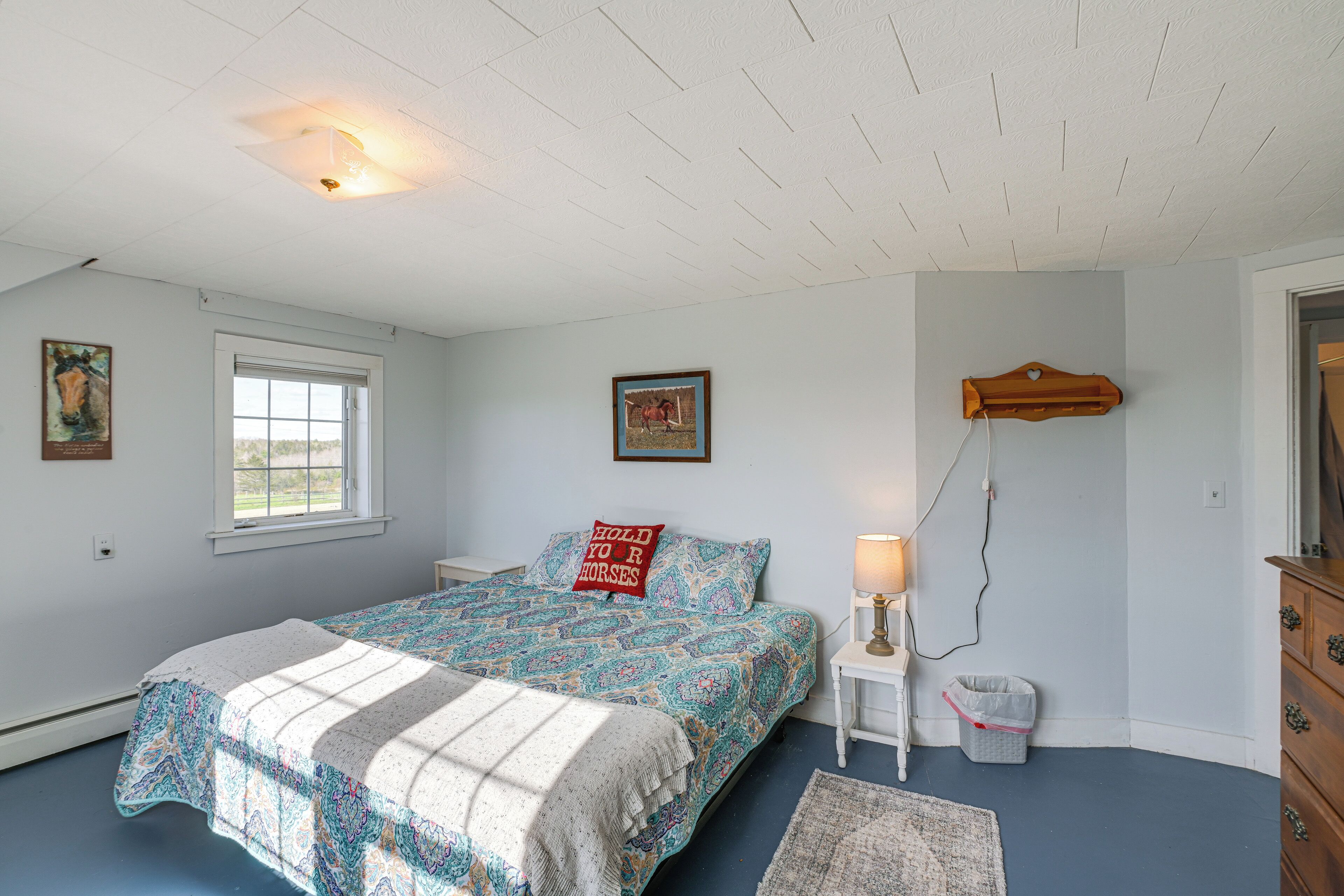 A bedroom with a bed, a window, a wall-mounted lamp, a dresser, and a trash bin.