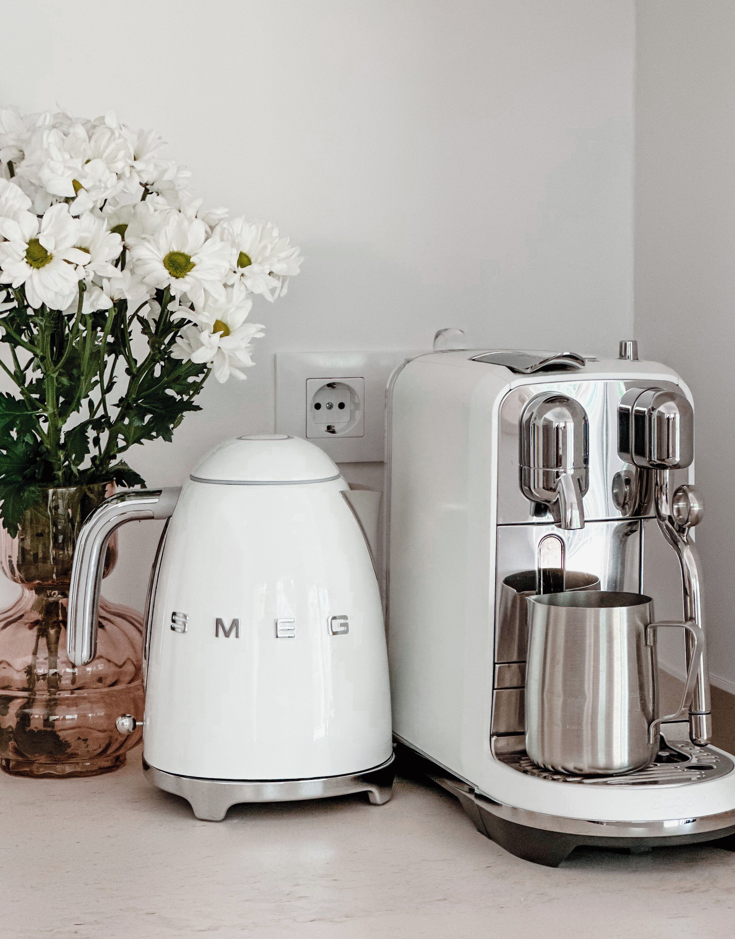A kitchen with a white electric kettle, a coffee maker, and a vase of white flowers on the counter.