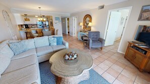 A spacious living room with a grey sofa, a round coffee table with decorative items, a grey armchair, and a television on a wooden stand.