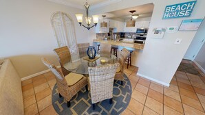 A dining area with wicker chairs and a glass table, a kitchen with white cabinets and modern appliances, and a 'Beach House' sign on the wall.