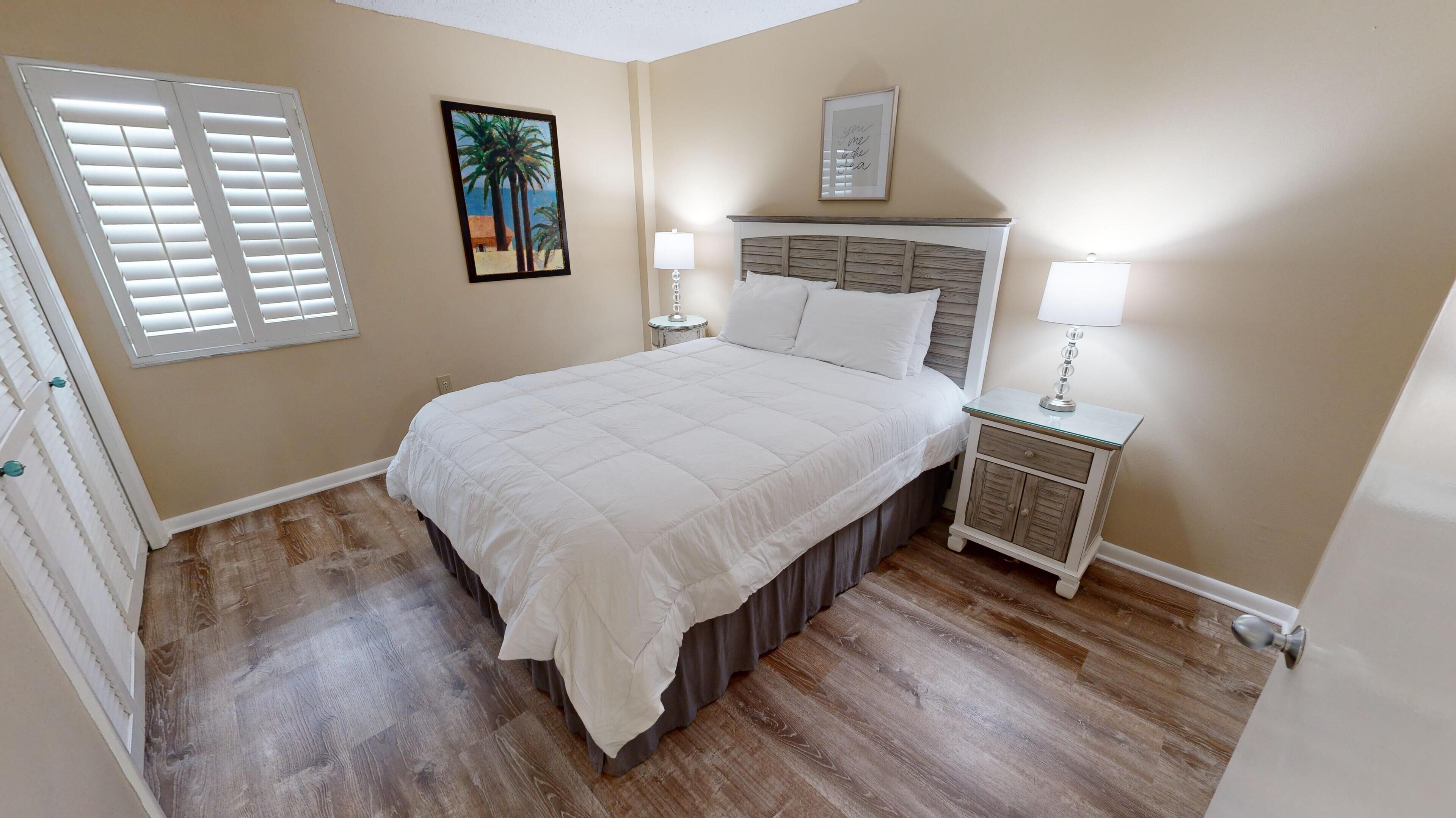 A bedroom with a large bed, a nightstand with a lamp, a framed picture on the wall, and a window with blinds.