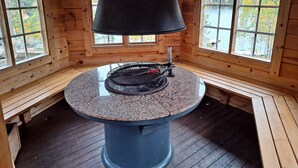 Interior d'una cabana de fusta amb una taula rodona de granat, una graella i finestres que donen vistes a un bosc i un llac.