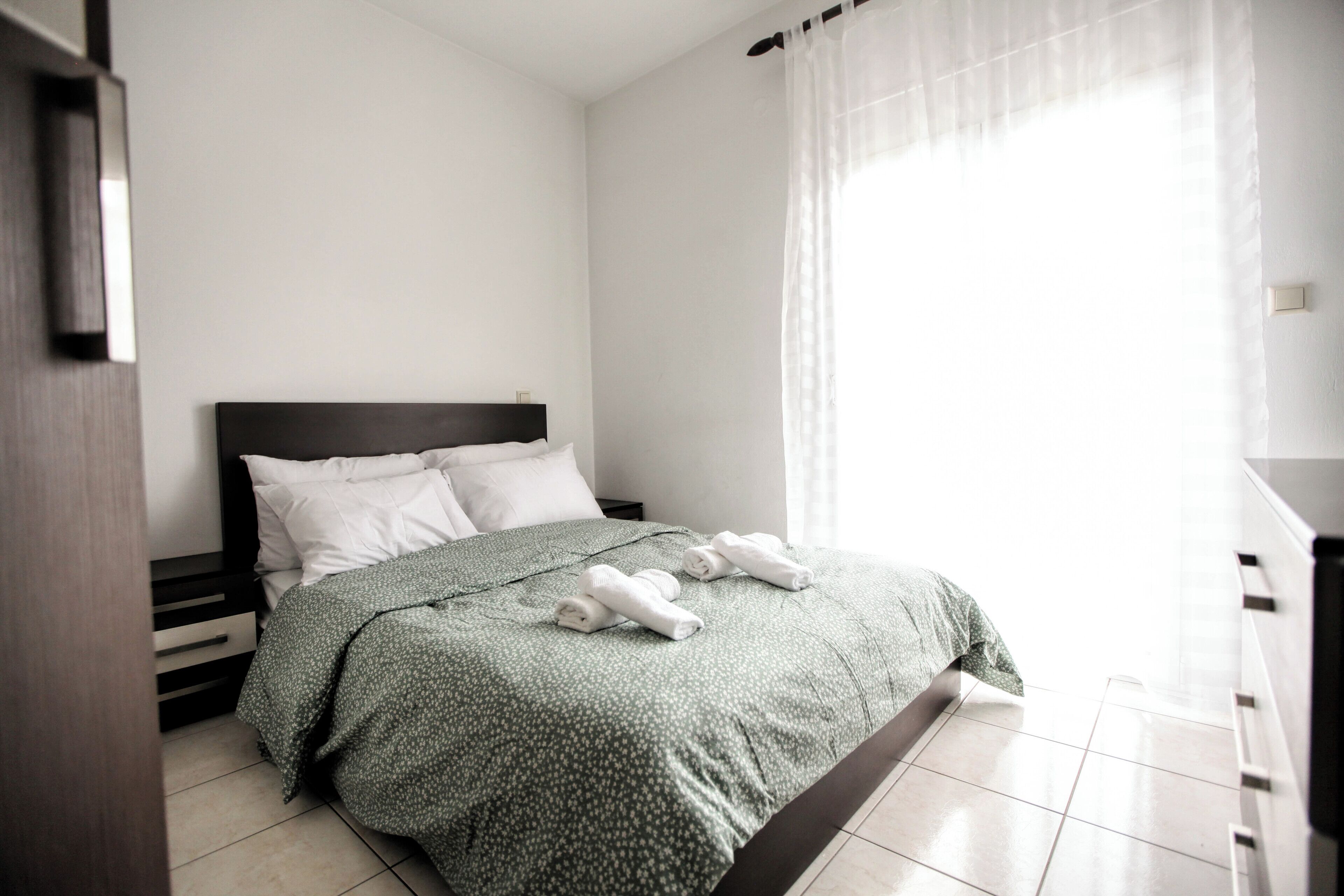 Un llit ben fet amb un edredó verd estampat i almoadines blanques, una nitrera de fusta amb una làmpada, i una finestra amb cortines de tela fina.