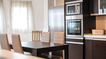 Una cocina moderna con horno empotrado, microondas y refrigerador. Hay una mesa de comedor con sillas y una ventana con cortinas.