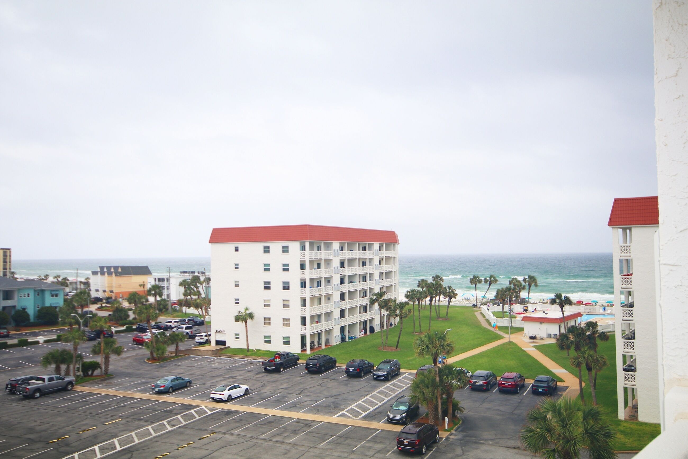 A multi-story hotel with a red roof, parking lot, and a view of the ocean.